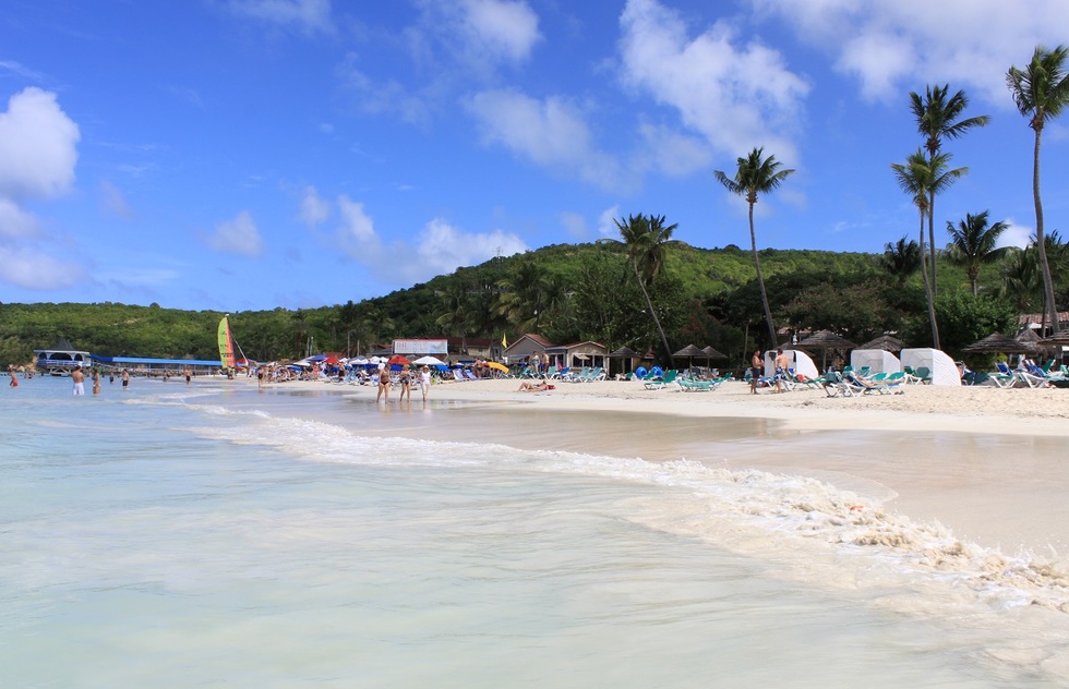 Tourists enjoying the beach at Dickenson Bay, Antigua, by standing in the water, lying on the sand, or walking along the shore.